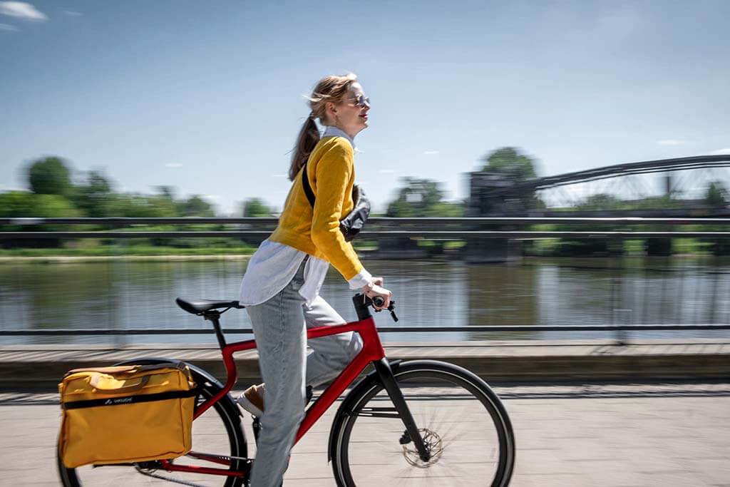Eine Frau fährt im Sommer auf einer Straße an einem Fluß entlang. Es symbolisiert das bequeme Fahren mit E-Bikes, in diesem Fall mit URWAHN Bikes