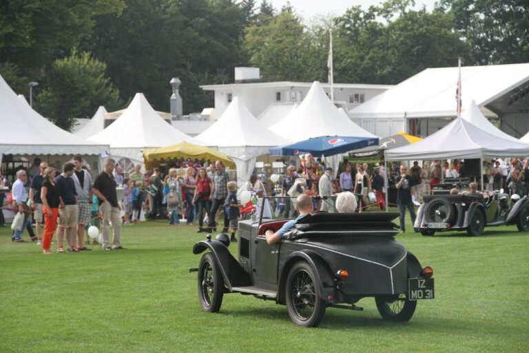 Ein britischer Oldtimer mit zwei Briten an Bord fährt über die Wiese. Im Hintergrund Festzelte vom British Flair 2026